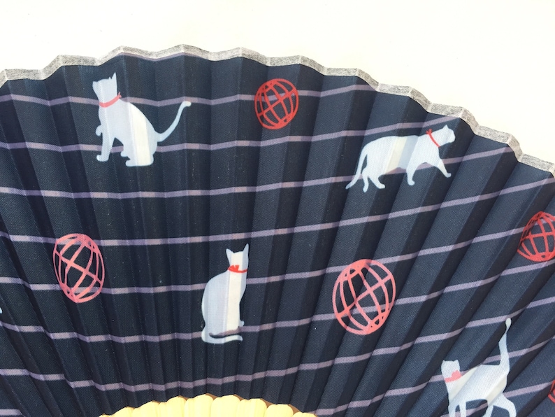 May include: A black folding fan with a white and red design. The fan features a repeating pattern of white cats and red globes.