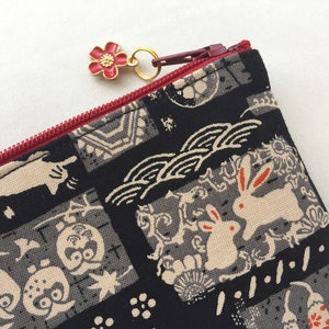 May include: A black zippered pouch with a red zipper and a red flower charm. The pouch is made of fabric with a Japanese-inspired pattern of rabbits, owls, and waves.