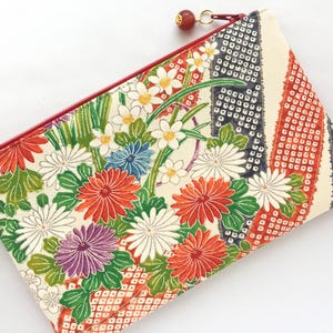 May include: A white zippered pouch with a floral pattern. The pouch features red, orange, white, green, and purple flowers on a white background. The zipper is red.