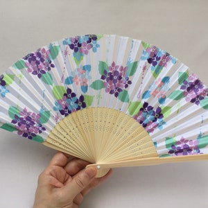 May include: A white folding fan with a floral design featuring purple, pink, and blue flowers. The fan is made of paper and has a wooden handle.