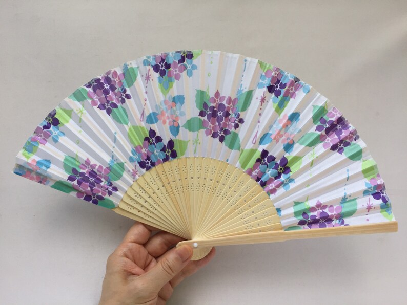 May include: A white folding fan with a floral design featuring purple, pink, and blue flowers. The fan is made of paper and has a wooden handle.
