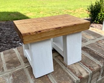 May include: A small wooden stool with a natural wood top and white painted legs. The stool is sitting on a brick surface.