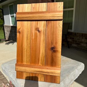May include: A rectangular wooden shutter with a natural finish. The shutter is constructed from vertical planks, with horizontal supports at the top and bottom. The wood grain and knots are visible, adding to its rustic charm. The shutter is designed for outdoor use.