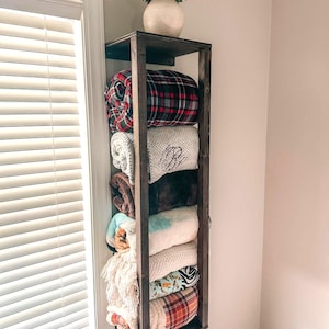 May include: A dark stained wooden blanket shelf mounted on a wall, filled with folded blankets in various colors and patterns. A white vase with greenery sits on top. The blankets include plaid, solid, and patterned designs.