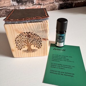 Könnte beinhalten: Eine Holzkiste mit einem geschnitzten Baumdesign und einem braunen Lederdeckel. Neben der Kiste steht eine kleine Flasche ätherisches Öl mit einem grünen Etikett, auf dem "Zirbenöl" steht. Das Etikett enthält auch Text über die Vorteile von Zirbenöl, darunter seine beruhigende Wirkung auf das Nervensystem und seine Fähigkeit, erholsamen Schlaf zu fördern.