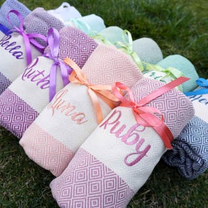 May include: Rolled personalized beach towels with embroidered names and diamond patterned borders. Each towel is tied with a satin ribbon in various colors, including purple, pink, and peach. The towels are arranged on green grass.