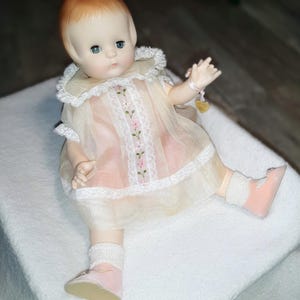 May include: A porcelain baby doll with red hair, blue eyes, and a pink dress with white lace trim and floral embroidery. The doll is wearing pink socks and shoes.