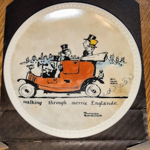 May include: A vintage ceramic plate with a black and white illustration of a red car with three people inside. The car is driving through a town with houses in the background. The text "walking through merrie Englande." is written below the car. The plate is signed "Norman Rockwell" in the bottom right corner.