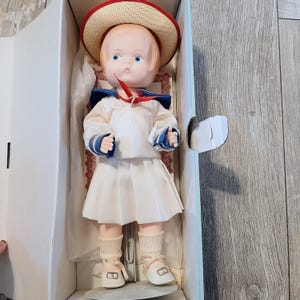 May include: A porcelain doll wearing a white sailor dress with a blue collar and red ribbon. The doll is wearing a straw hat with a red band and white socks and shoes. The doll is in a white box with a red and white patterned paper.