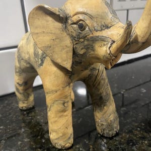 May include: A carved wooden elephant figurine with a light brown and dark grey marbled pattern. The elephant has large ears, a trunk, and four legs. The figurine is a decorative item.