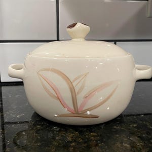 May include: Cream-colored ceramic soup tureen with a lid and two handles. The tureen is decorated with a pink and brown leaf design. The lid has a small brown knob. The tureen is sitting on a dark surface.