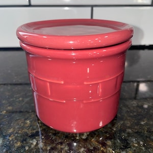 May include: A red ceramic jar with a lid. The jar has a textured design around the middle and sits on a dark speckled surface. The lid is a matching red color and fits snugly on top.