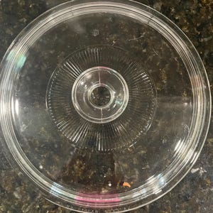 May include: A circular, clear glass lid featuring a central knob handle. The lid has a series of radiating, linear ridges extending from the center toward the outer edge, with a smooth, raised rim around the perimeter and a small pink label.