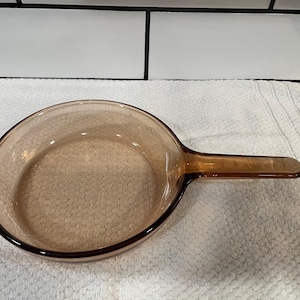 May include: A clear, amber-colored glass skillet with a long handle. The skillet has a round cooking surface and a dark brown rim. The handle is also amber-colored and extends from the side of the pan.