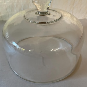 May include: A clear glass dome cover with a smooth, rounded top and a small, solid glass knob handle centered on the top. The dome has a wide, circular base and a transparent, polished finish, designed to fit over food or decorative items.
