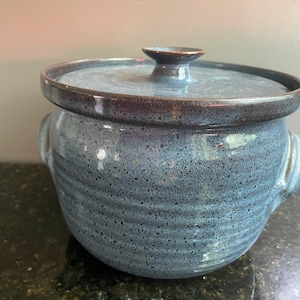 May include: A round, blue ceramic pot with a speckled finish and horizontal ridges along the body. The pot features two side handles and a matching lid with a central knob. The glaze has a mottled appearance with darker blue and grey tones.
