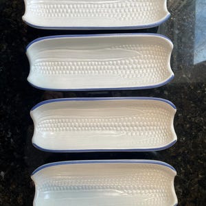 May include: Four white ceramic corn-shaped serving dishes with a blue rim. The dishes have a textured surface resembling corn kernels. The dishes are arranged in a rectangular pattern, perfect for serving corn on the cob or other side dishes.