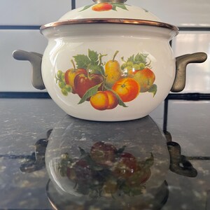 May include: A white enamel pot with a lid, featuring a fruit design of apples, pears, grapes, and peaches. The pot has bronze-colored handles and rim. The lid is topped with a bronze knob. A vintage kitchenware item.