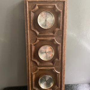 May include: A rectangular brown wood-grain wall-mounted weather station featuring three circular brass-rimmed gauges. The top gauge is a thermometer, the middle is a barometer, and the bottom is a hygrometer. A decorative metal ring is at the top.
