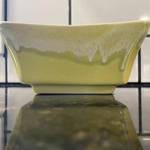 May include: A rectangular, light green ceramic bowl with a white, dripped glaze effect on the rim. The bowl is sitting on a reflective surface, with a white tile background.
