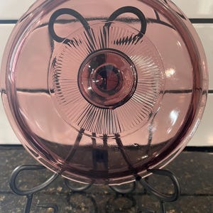 May include: A vintage, round, pink glass lid with a central design and radial lines, resting on a black metal stand. The lid has a slightly domed shape and a dark center. The stand has a decorative, curved design.