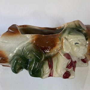 May include: A rectangular ceramic planter with a whimsical dog design. The planter features a brown, white, and green color scheme with red accents. The dog's face is visible on the side, adding a playful touch to this decorative item.