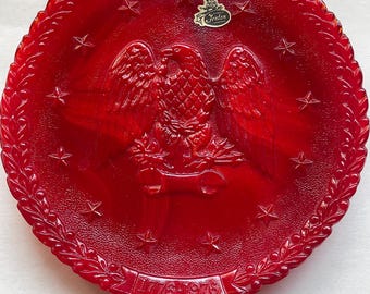 May include: A round red glass plate featuring a central embossed eagle design, surrounded by a ring of stars and a laurel leaf border. The text 1776-1976 is embossed at the bottom, and a Fenton sticker is attached to the upper surface.