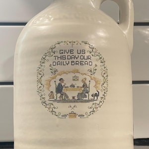 May include: A white ceramic pitcher with a handle and a cross-stitch design that says "Give us this day our daily bread". The design features a couple sitting at a table with a cat.