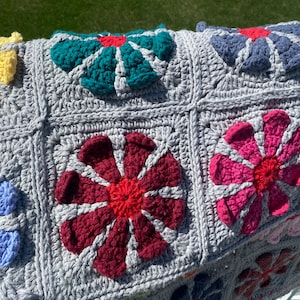 Puede incluir: Una manta de crochet gris con motivos de flores de colores. Las flores est&aacute;n hechas en varios colores, incluyendo rojo, rosa, azul, amarillo y verde.
