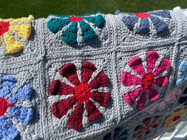 Puede incluir: Una manta de crochet gris con motivos de flores de colores. Las flores est&aacute;n hechas en varios colores, incluyendo rojo, rosa, azul, amarillo y verde.