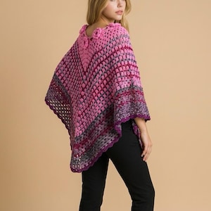 May include: A pink and purple crochet poncho with a floral neckline. The poncho has a geometric pattern with a dark purple trim. The model is wearing black pants.
