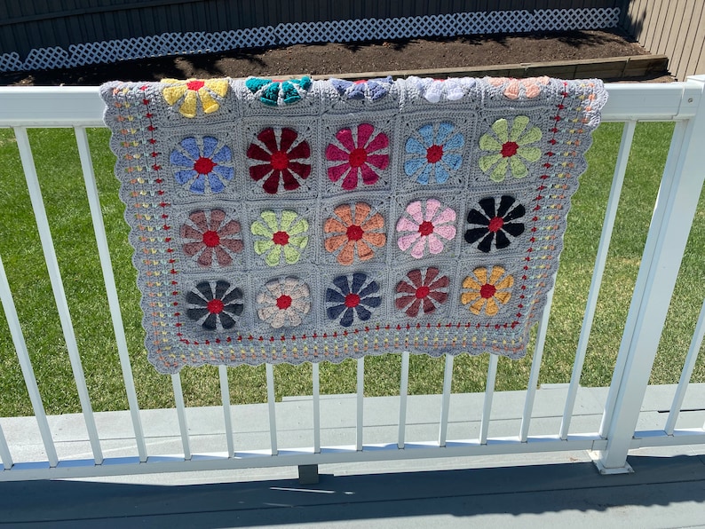 Puede incluir: Una manta de ganchillo con fondo gris y dise&ntilde;os de flores de colores. La manta est&aacute; colgada en una barandilla blanca.