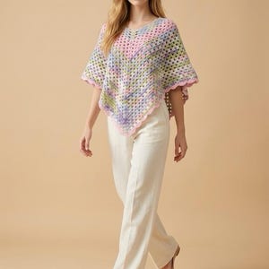 May include: A colorful crochet poncho in pastel shades of pink, blue, green, and purple. The poncho has a V-neck and scalloped edges, paired with white pants and beige flats. The overall style is bohemian and handmade.