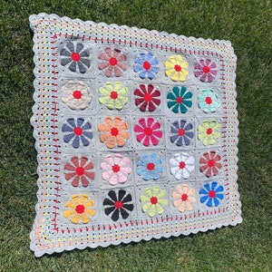 Puede incluir: Una manta de crochet gris con un borde de hilo rojo, amarillo y naranja. La manta est&aacute; hecha de cuadrados, cada uno con un dise&ntilde;o de flor colorido en el centro.