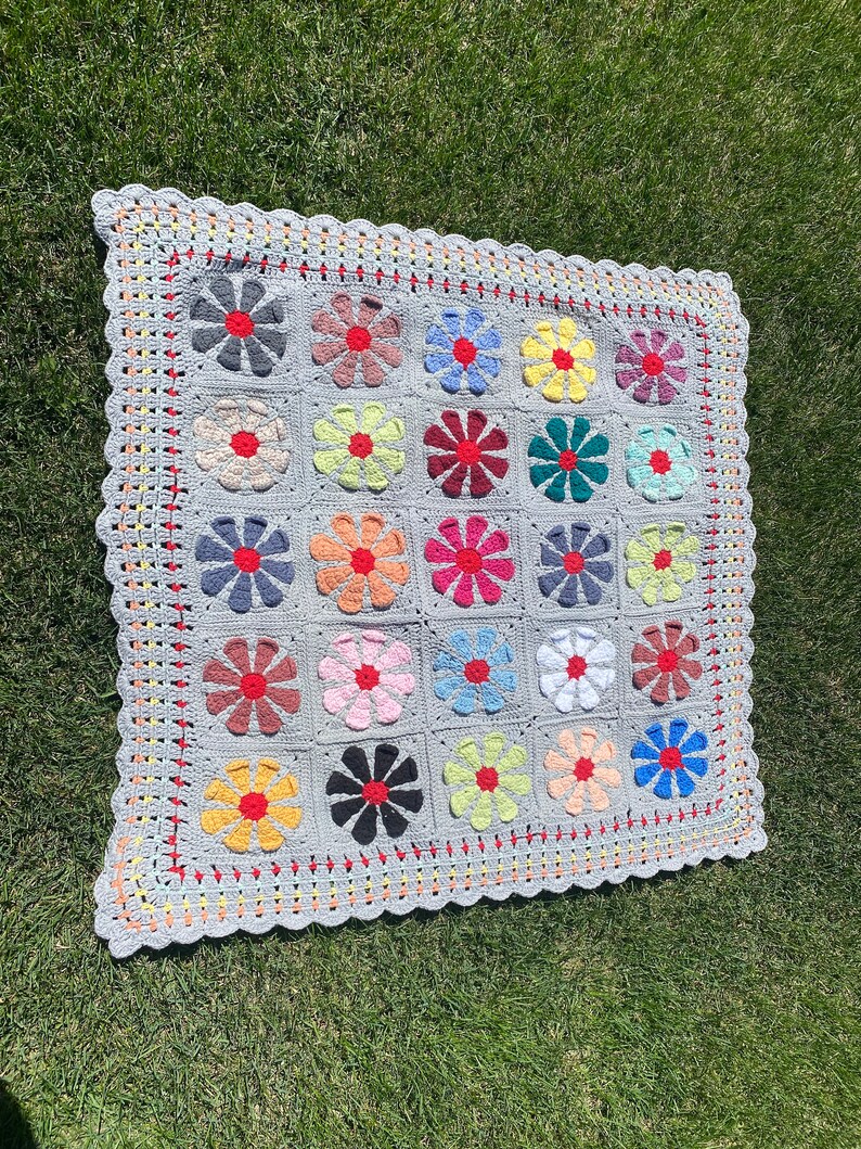 Puede incluir: Una manta de crochet gris con un borde de hilo rojo, amarillo y naranja. La manta est&aacute; hecha de cuadrados, cada uno con un dise&ntilde;o de flor colorido en el centro.