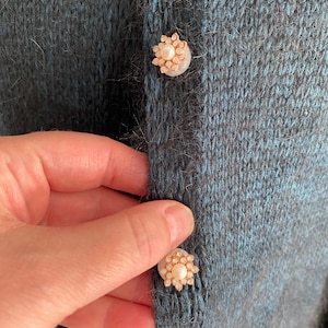 May include: A close-up of a dark blue knitted cardigan with two pearl and gold buttons. The buttons are in the shape of a flower.