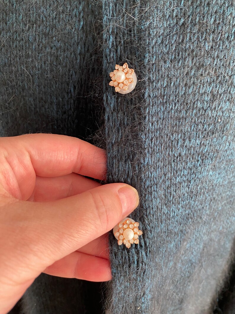 May include: A close-up of a dark blue knitted cardigan with two pearl and gold buttons. The buttons are in the shape of a flower.