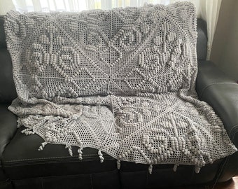 Puede incluir: Una manta de ganchillo gris con un patrón texturizado en relieve. La manta está drapeada sobre un sofá gris.