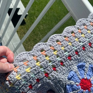 Puede incluir: Una manta de ganchillo gris con un borde festoneado y detalles coloridos. La manta presenta un gran dise&ntilde;o floral en tonos de azul, rojo y blanco.