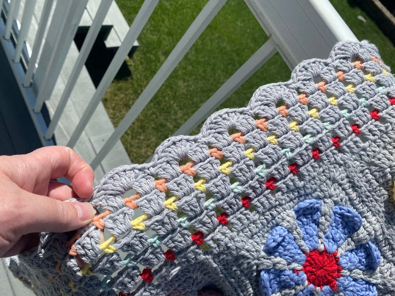Puede incluir: Una manta de ganchillo gris con un borde festoneado y detalles coloridos. La manta presenta un gran dise&ntilde;o floral en tonos de azul, rojo y blanco.
