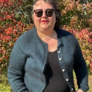 May include: A woman wearing a teal blue cardigan with pearl buttons over a black top. She is standing in front of a tree with red leaves.