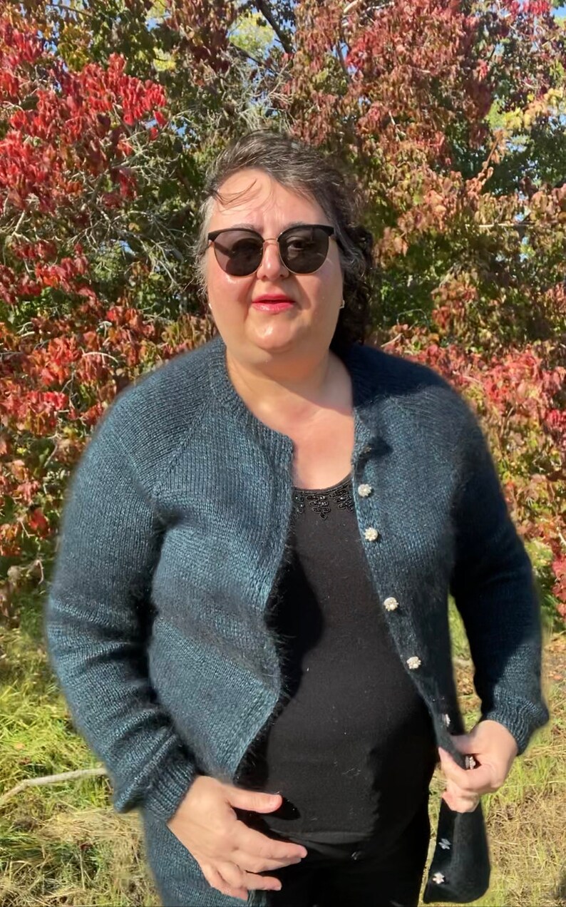 May include: A woman wearing a teal blue cardigan with pearl buttons over a black top. She is standing in front of a tree with red leaves.
