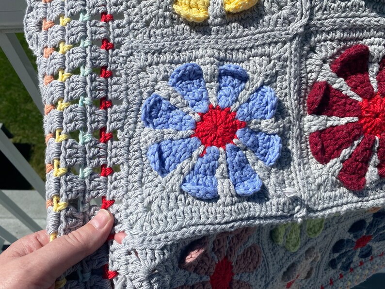 Puede incluir: Una manta de ganchillo con fondo gris y dise&ntilde;os de flores de colores. La manta est&aacute; hecha con hilo de diferentes colores y tiene una superficie texturizada.