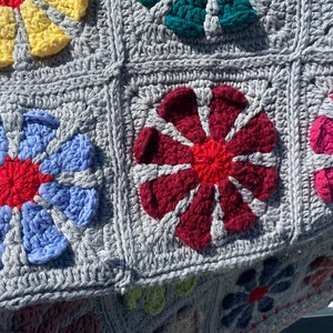 Puede incluir: Una manta de ganchillo con fondo gris y cuadrados de flores de colores. Las flores son en tonos de azul, rojo, rosa, amarillo y verde.
