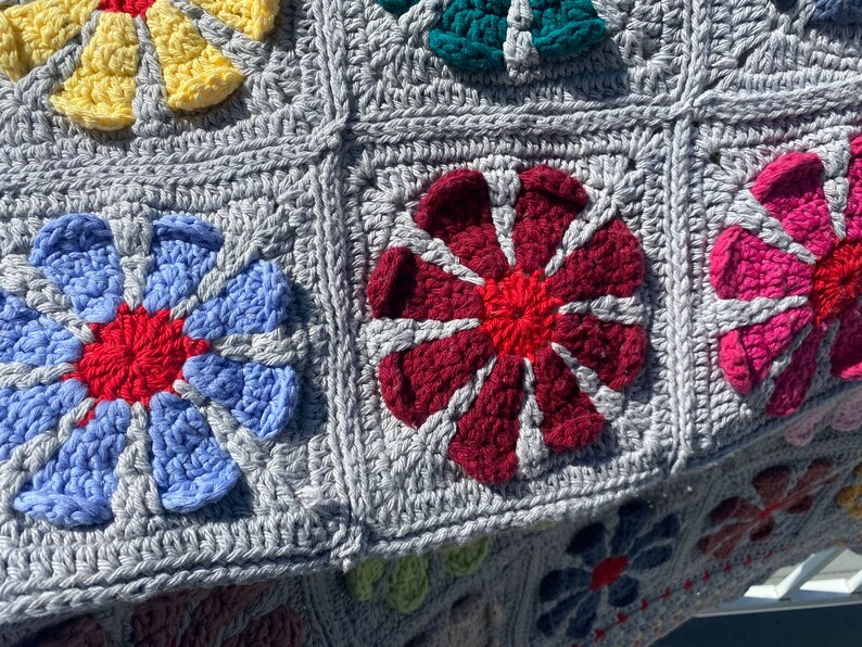 Puede incluir: Una manta de ganchillo con fondo gris y cuadrados de flores de colores. Las flores son en tonos de azul, rojo, rosa, amarillo y verde.