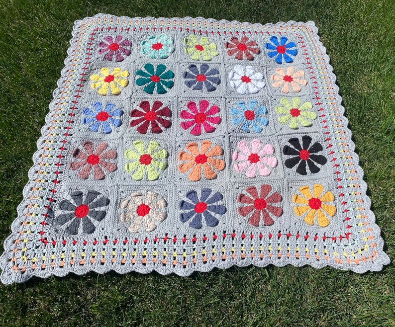 Puede incluir: Una manta de ganchillo con fondo gris y dise&ntilde;os de flores de colores. La manta tiene un borde festoneado y est&aacute; hecha con una variedad de hilos de colores diferentes.