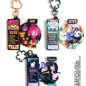 May include: A collection of colorful fanmade keychains featuring anime-style artwork. The keychains showcase various characters and include text such as "STOMACH BOOK", "KIKUO", and "ADO TOP HITS". Each keychain has a unique design and a metal clasp.