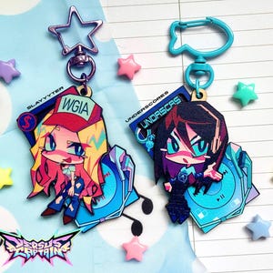 May include: Two acrylic keychains featuring anime-style characters. The left keychain has a purple star clasp and a character with blonde hair wearing a red cap labeled WGIA. The right keychain has a blue clasp and a character with dark hair labeled UNDERSCORES. Both feature blue music player graphics.