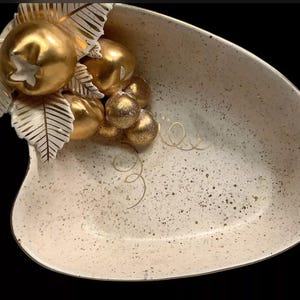 May include: Cream-colored, speckled ceramic serving dish with a gold-painted fruit and leaf cluster. The dish has a unique, asymmetrical shape and is suitable for serving appetizers or decorative display.