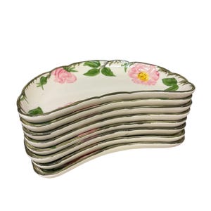 May include: A stack of six crescent-shaped ceramic dishes with a cream-colored base. Each dish features a hand-painted floral motif of pink roses and green leaves along the curved edge, finished with a dark green painted rim.